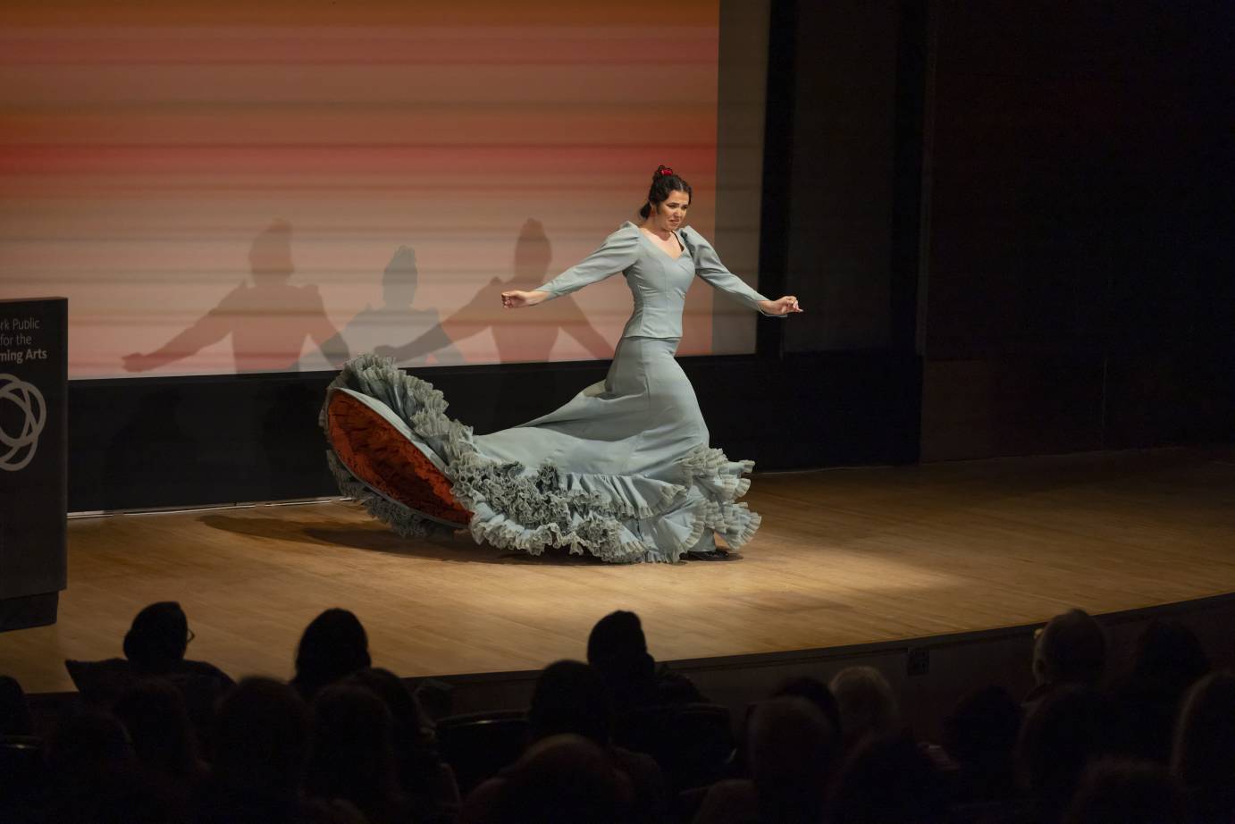 A flamenco dancer in a light aqua dress, holding castanets near her sides, flips her ruffled skirt to show the red beneath. 